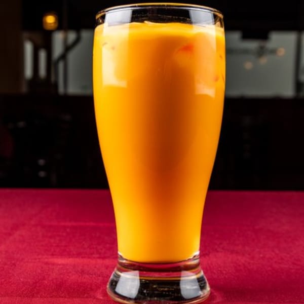 A tall glass of vibrant orange mango lassi with ice, on a red tablecloth.