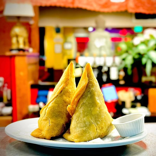 Two golden-brown Indian samosas and a small cup of sauce on a white plate in a restaurant.
